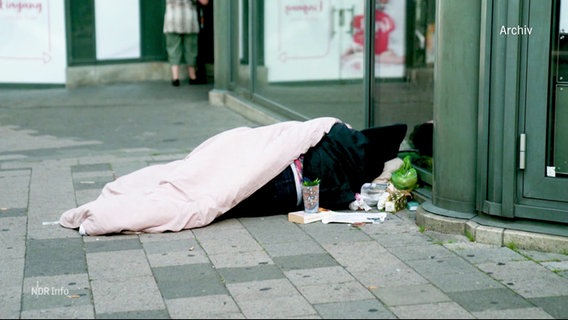 Ein obdachloser Mensch liegt auf der Straße an einer Häuserwand. © Screenshot 