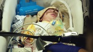 Ein Kleinkind ist an medizinische Geräte angeschlossen und liegt in einem Transport-Körbchen. © Screenshot 