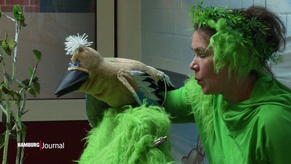 Szene aus dem Stück "Oh Oma O" vom Theater Zeppelin: Eine Schauspielerin in grünem Kostüm mit einem Vogel als Handpuppe. © Screenshot 