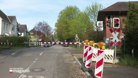 Ein Bahnübergang an einer Straße, von rot-weißen Schranken gesperrt. Rechts im Vordergrund Warnbaken einer Baustelle. © Screenshot 