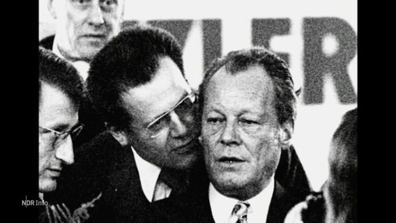 Eine schwarz-weiß Aufnahme von Willy Brandt und Günter Guillaume: Guillaume flüstert Brandt etwas ins Ohr. © Screenshot 
