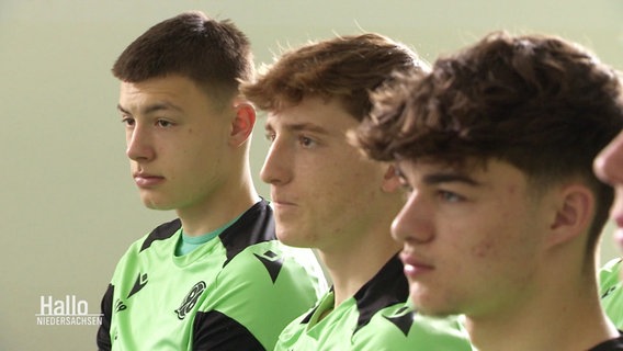 Drei junge Fußballspieler sitzen in hellgrünen Trikots nebeneinander. © Screenshot 