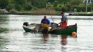 Zwei Fischer stehen in ihrem Boot und holen ein Netz ein. © Screenshot 