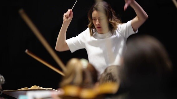 Eine junge Frau dirigiert ein Orchester. Sie hebt Hände und Taktstock in einer dynamischen Geste. © Screenshot 