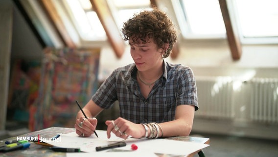 Ein junger Student sitzt an einem Tisch und malt. © Screenshot 