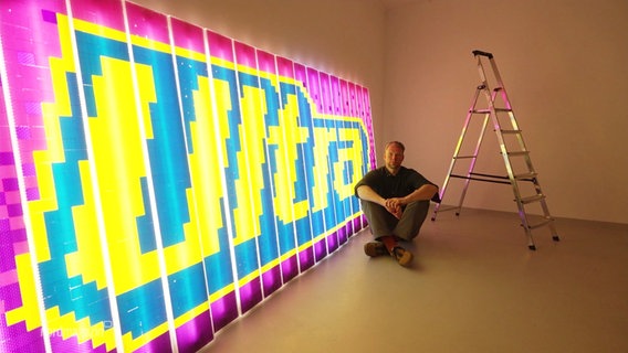 Künstler Tino Bittner mit einem leuchtenden Ausstellungsstück welches das Wort "Ultra" formt. © Screenshot 