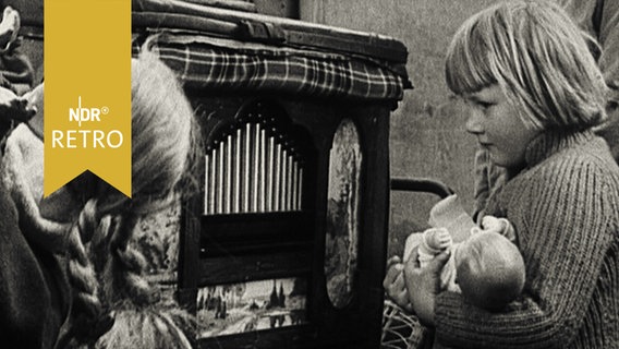 Mädchen mit Puppe im Arm steht fasziniert vor einer Drehorgel (1955)  