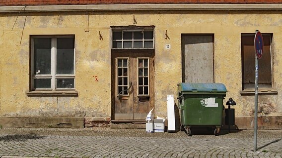 Heruntergekommene Fassade eines leerstehenden Hauses. Zwei von drei Fenstern sind mit Brettern verschlossen. Davor stehen ein großer Müllcontainer sowie Kartons. © MDR/Hoferichter & Jacobs 