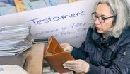 Monate: Rechtsanwältin beim Durchblättern von Unterlagen; im Hintergrund Geldscheine und ein Schriftstück mit dem Wort Testament. © NDR 