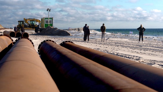 Auf einem Strand von Sylt lagern große Rohre, die für Sandaufspülungen verwendet werden. Im Hintergrund sind ein Bagger und ein Bauhäuschen zu erkennen. Ein paar Menschen stehen am Strand und schauen aufs Meer. © NDR 