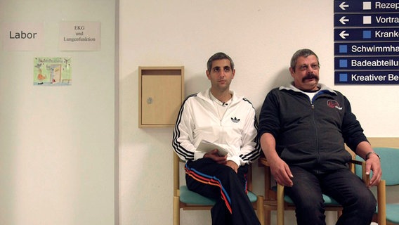 Michel Abdollahi (l.) sitzt mit einem Mann vor einer Tür in einer Rehaklinik. © NDR 