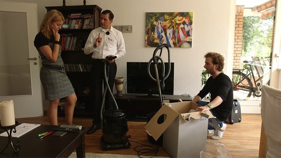 Zwei Männer zeigen einer Frau in ihrem Wohnzimmer einen neuen Staubsauger.  
