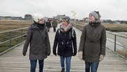Drei junge Frauen gehen in Winterjacken und mit Mützen einen Steg am Strand entlang. © NDR/7Tage 