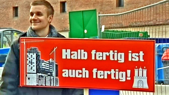 Tobias Schlegl steht vor der Elbphilharmonie und hält ein Schild mit der Aufschrift "Halb fertig ist auch fertig!" in der Hand.  