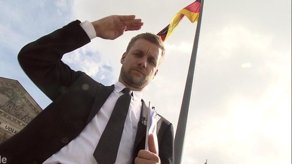 Tobias Schlegel vor einer Deutschland-Fahne erhebt die Hand zum Gruß  