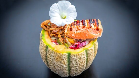 Melonensalat und Lachs in eine halbe Melone gefüllt und mit Blüten dekoriert angerichtet auf einem Teller. © NDR Foto: Dave Hänsel