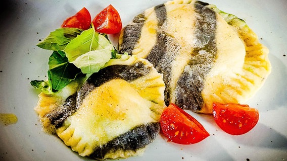 Ein Teller mit gelb-schwarzen Ravioli und Tomaten. © NDR Foto: Dave Hänsel