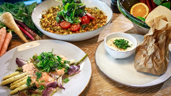 Auf einem Tisch stehen ein Teller mit Linsensalat, Spargel und Brokkoli mit Lachsforelle sowie Quarkcreme und Rhabarber in einem Pergament-Päckchen. © NDR Foto: Tarik Rose