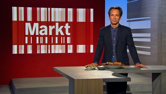Der Moderator Jo Hiller steht an seinem Pult, vor ihm ist Essen aufgebaut. Im Hintergrund ist das Markt-Logo zu sehen. © NDR 