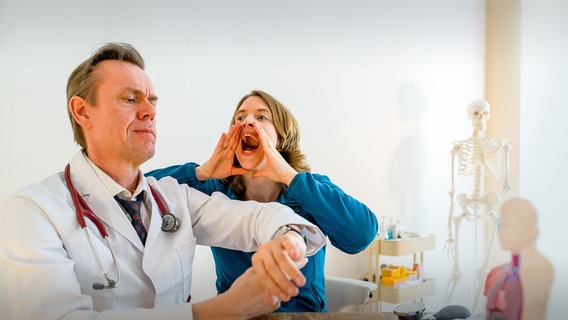 Karikaturistische Szene: Arzt in Kittel schaut auf die Uhr, Frau daneben hat die Hände an den Mund gelegt und versucht sich laut Gehör zu schaffen. © SWR 