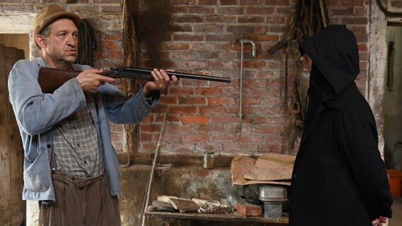 Szenenbild der 92. Büttenwarder-Folge "Der Tod ist ein sturer Arsch": Ein Mann hält ein Gewehr auf eine Gestalt gerichtet. © NDR/Nico Maack 
