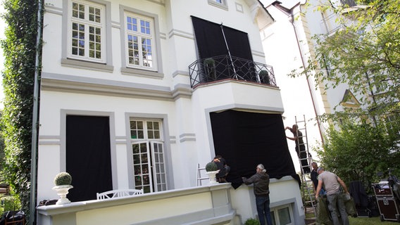 Die Fenster einer weißen Villa werden von vier Lichttechnikern mit schwarzem Molton abgeklebt. © NDR Foto: Claudia Timmann