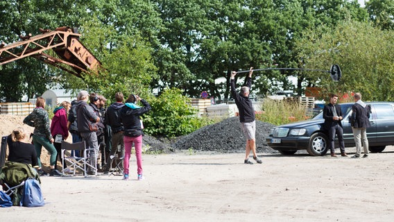 In einer Kiesgrube steht links das Filmteam, rechts stehen zwei Schauspieler vor einem schwarzen Auto. Der Tontechniker steht in der Mitte mit einer Tonangel. © NDR Foto: Claudia Timmann
