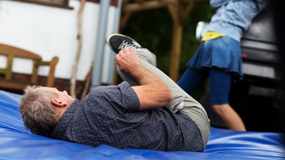 Stucke (Michael Lott) liegt auf einer blauen Matte.  Im Hintergrund steht Ceyda (Merle de Villiers) mit ausgestrecktem Bein. © NDR Foto: Claudia Timmann