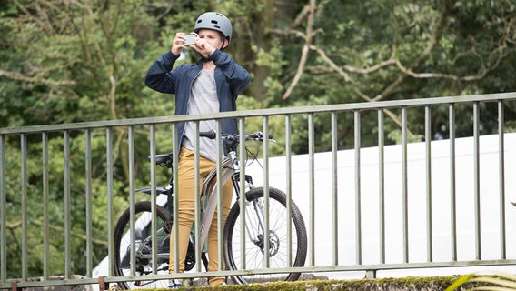 Johannes (Luke Matt Röntgen) steht mit seinem Fahrrad auf einer Brücke und fotografiert mit seinem Handy. © NDR Foto: Claudia Timmann