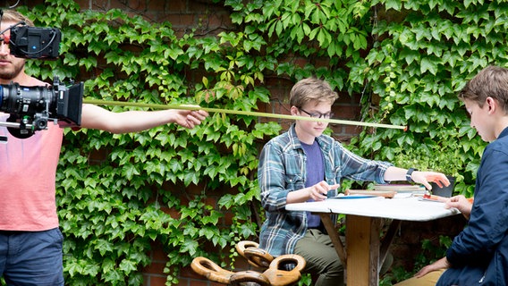 Benny (Ruben Storck) und Johannes (Luke Matt Röntgen) sitzen an einem Tisch. Der Kameramann hält ein Maßband. © NDR Foto: Claudia Timmann