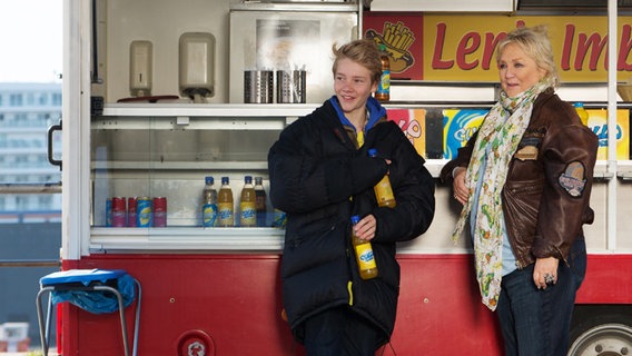 Max und Oma Leni stehen vor dem Imbisswagen. Max hat eine dike Jacke an und hält zwei Flaschen Limonade. © NDR Foto: Claudia Timmann