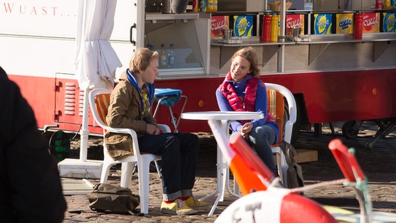 Max und Tatja sitzen an einem Tisch am Hafen vor Lenis Imbiss. © NDR Foto: Claudia Timmann
