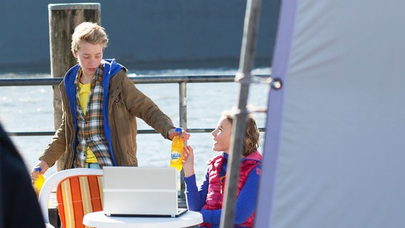 Tatja sitzt an einem Tisch am Hafen. Max steht daneben und reicht ihr eine Flasche Limonade. © NDR Foto: Claudia Timmann