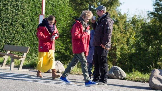 Nina und Max gehen in warmen Jacken mit einem Becher in der Hand auf einer Straße. Zwei Menschen stehen im Hintergrund. © NDR Foto: Claudia Timmann