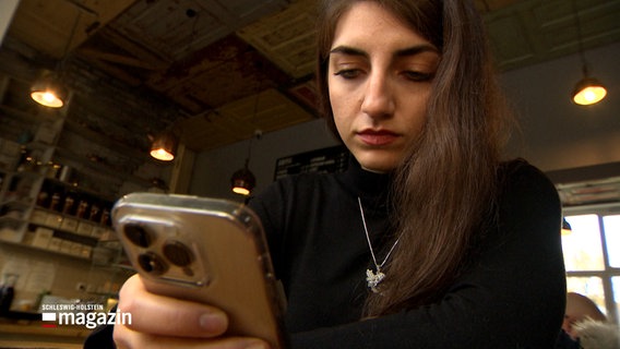 Eine junge Frau sitzt an einem Tisch in einem Cafe und blickt auf ihr Smartphone. © NDR 