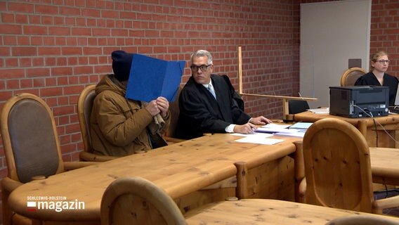 Ein Angeklagter sitzt in einem Gerichtssaal und spricht mit seinem Verteidiger und schirmt sein Gesicht mit einer blauen Mappe ab. © NDR 