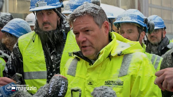 Bundeswirtschaftsminister Robert Habeck (B.90/Grüne) steht mit anderen Menschen auf dem Gelände eine Betonwerks in Norwegen und spricht mit zahlreichen Journalisten während es schneit. © ARD 