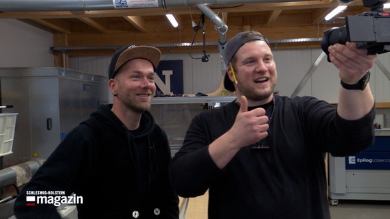 Zwei junge Handwerker stehen in einer Werkstatt und lächeln in die Kamera, die der Handwerker rechts im Bild am Objektiv hochhält. © NDR 