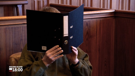 Ein Angeklagter sitzt in einem Gerichtssaal und hält einen aufgeklappten Aktenordner vor seinem Gesicht hoch. © NDR 