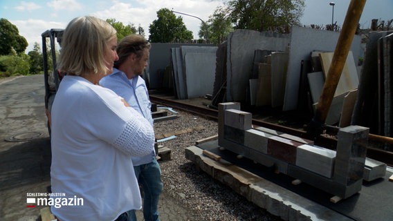 Ein Mann und eine Frau betrachten eine Wand aus Grabsteinen. Die Steine sind zum Gedenken an verstorbene Wohnungslose. © NDR Foto: NDR Screenshots