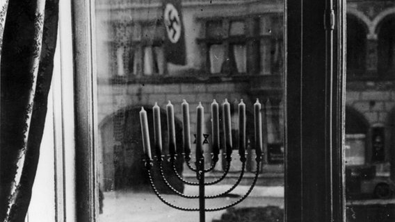 Ein schwarz-Weiß Foto zeigt einen Chanukka-Leuchter vor einem Fenster auf der Straße gegenüber ist die NSDAP-Zentrale. © NDR Foto: Rosi Rachel Posner
