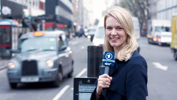 ARD Korrespondentin Julie Kurz steht an einer Fußgängerampel und spricht in ein Mikrofon.  