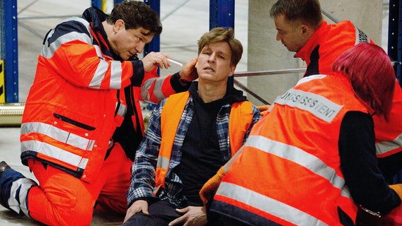 Christian (Francis Fulton-Smith, l.) ist als Notarzt vor Ort: Boris Kehr (Jürgen Klein, M.) wurde von einer herabfallenden Pallette verletzt (mit Komparsen, r.). © NDR/ARD/Volker Roloff 