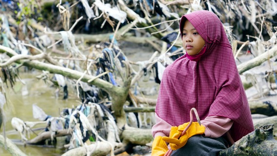 Ein indonesisches Mädchen sitzt an einem Gewässer, das mit Plastik verschmutzt ist. © BR/WDR/Yetha/a&o büro 