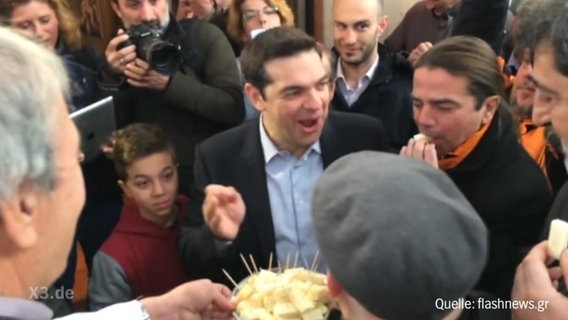 Tsipras auf einer Veranstaltung mit einem Käsaespieß in der Hand.  
