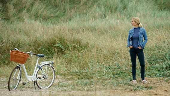 Szenenbild aus der dänischen Serie "Whits Sands": Eine Frau steht in den Dünen, neben ihr ein weißes Fahrrad. © NDR/TV2. Deluca Films Foto: Martin Dam Kristensen