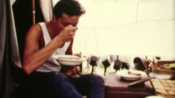 Ein junger Mann nimmt in seinem Zelt in den 50er-/60er-Jahren eine Mahlzeit zu sich. © NDR 