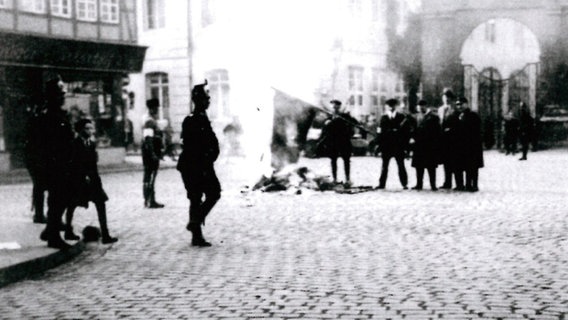 Menschen stehen um ein Feuer auf dem Ackerhof vor dem Braunschweiger Volksfreund-Haus, ein Mann hält eine große Fahne in die Flammen. Menschen stehen um ein Feuer auf dem Ackerhof vor dem Braunschweiger Volksfreund-Haus, ein Mann hält eine große Fahne in die Flammen. © Walter Schmidt/Nachlass Archiv IBRG