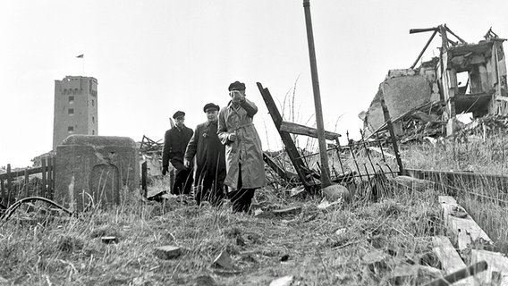 Drei Bewohner besichtigen am 1. März 1952 die völlig zerstörte Hauptstraße der Insel Helgoland. © dpa Foto: Jochen Blume