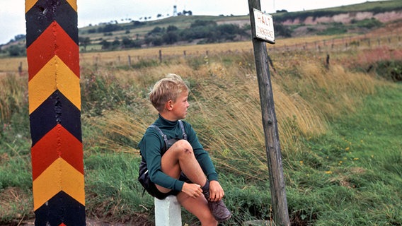 Ein Junge sitzt auf einem weißen Pfosten zwischen einem Grenzpfahl mit dem Wappen der DDR und einem Schild mit der Aufschrift "Halt Zonengrenze". © picture-alliance / dpa Foto: Wilhelm Bertram